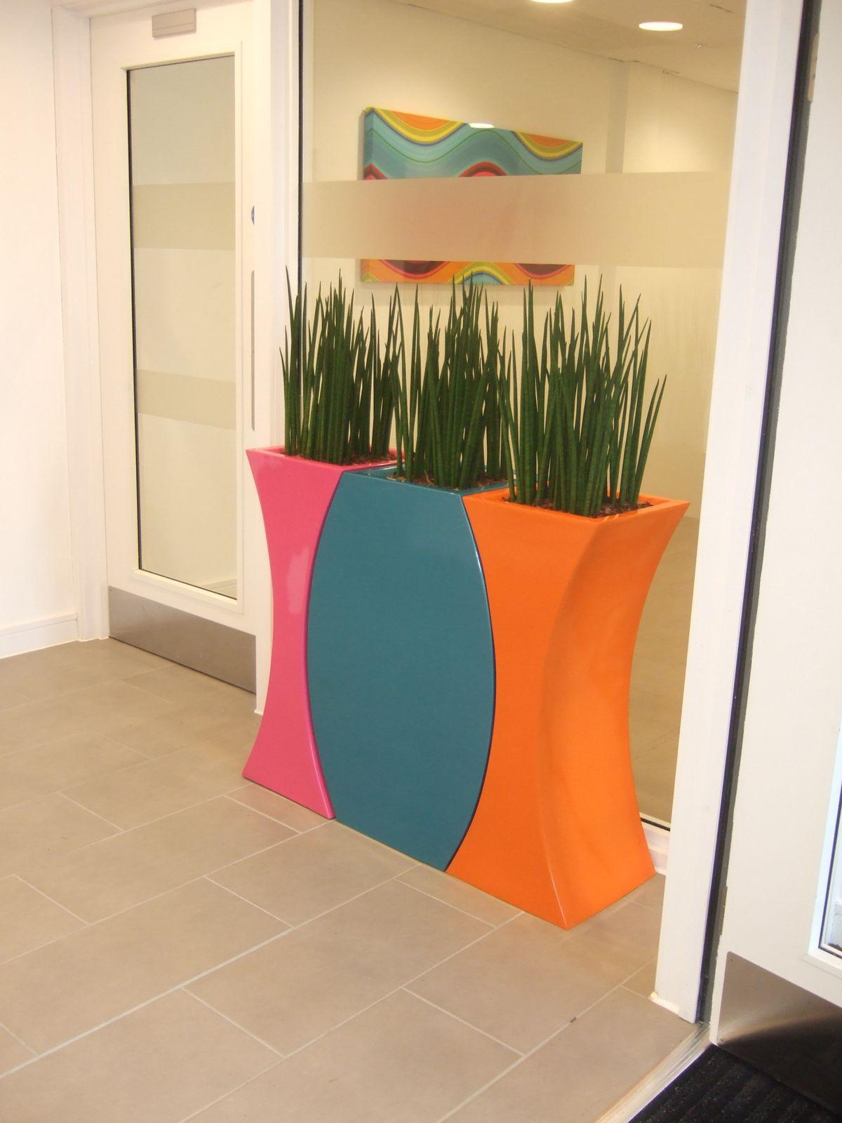 A modern planter with tall green plants stands in a hallway. The planter is colorful with orange, pink, and blue curved panels. A frosted glass door and a colorful abstract painting are in the background.