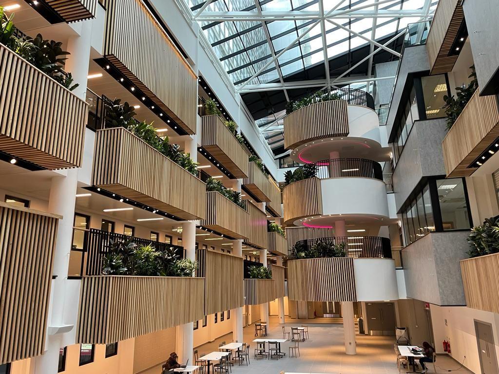 Modern atrium with multiple floors, featuring wood paneling, glass railings, indoor plants, and abundant natural light from a large glass ceiling. Tables and chairs are arranged on the ground floor.