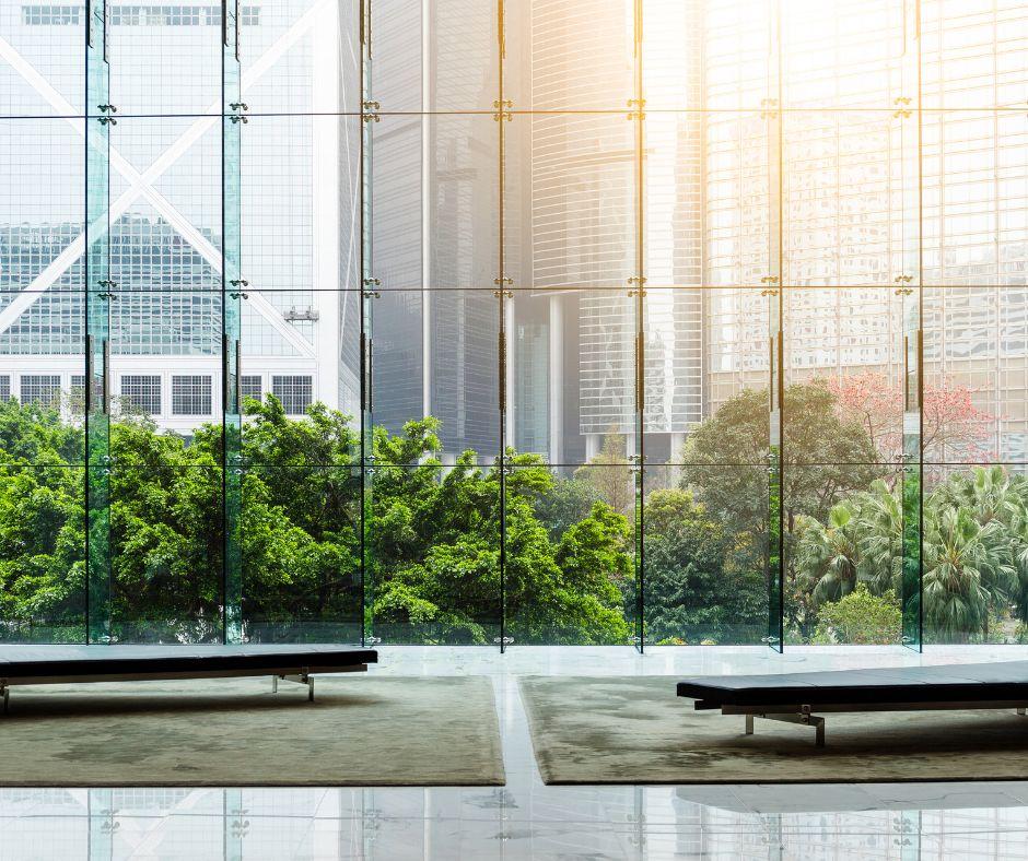 A modern lobby with large floor-to-ceiling glass windows overlooks green trees and tall city buildings, letting in plenty of natural light. Two simple benches are placed on a shiny floor.