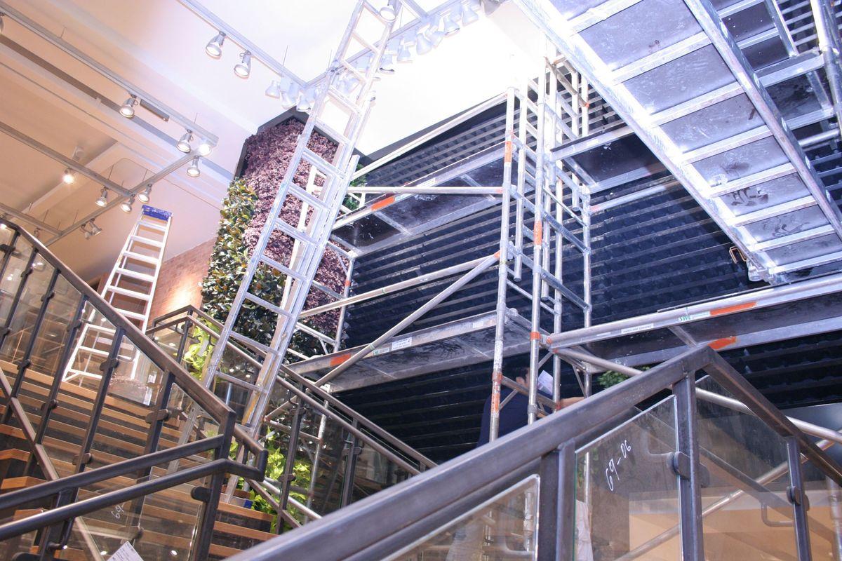 Metal scaffolding and ladders are set up inside a modern building with glass railings and stairs, positioned near a large green wall installation by Urban Planters covered with plants and industrial lighting overhead.