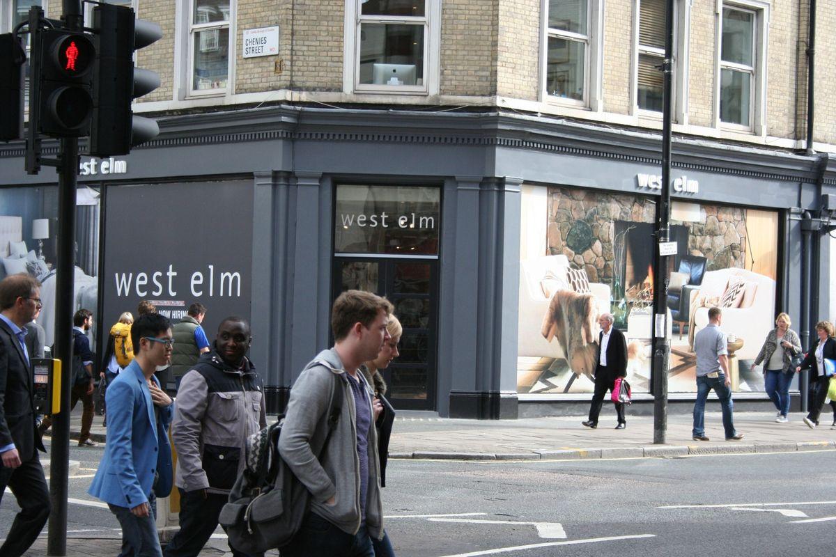 People cross a city street at a crosswalk in front of a West Elm store on a corner. The traffic light is red, and some pedestrians walk while others wait near the sidewalk.