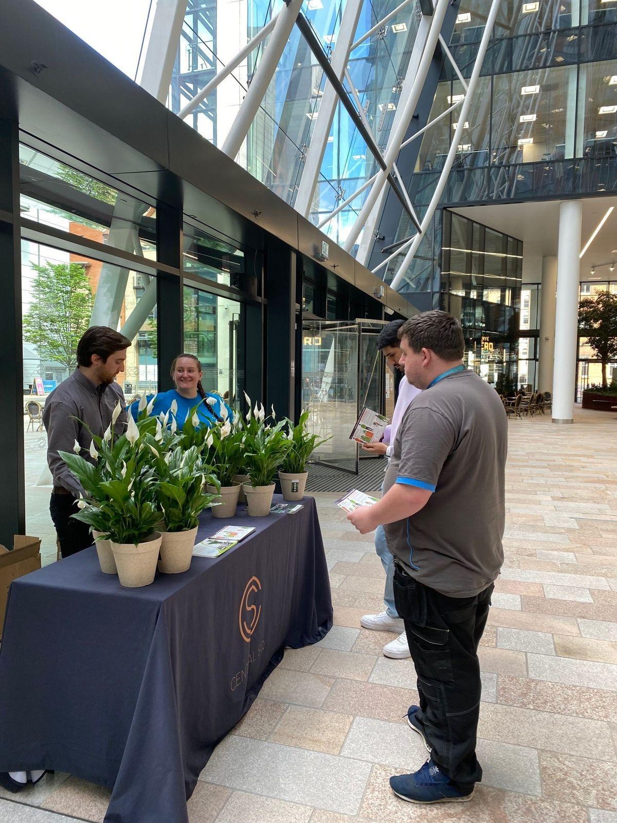 Urban Planters team stand near a table with potted plants inside a modern glass building; two are behind the table, talking to two others who are holding papers.