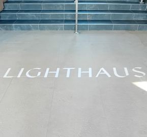 Sleek light gray stone entrance floor displaying 'LIGHTHAUS' in large white block lettering leading up to a set of dark slate tiled stairs with a central metal handrail