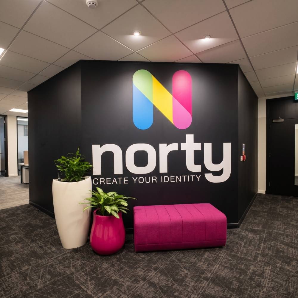A stylish office reception area featuring a black feature wall with the colorful Norty logo, a bright pink bench, and potted plants.