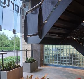 A modern steel staircase viewed from below near large glass windows, with concrete planter boxes lining the floor beneath and a glass-block wall in the background.