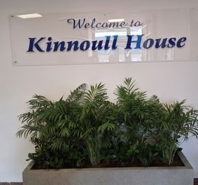 Acrylic sign reading Welcome to Kinnoull House above a large planter with leafy green plants in a building lobby.