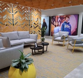 A spacious office seating area featuring grey sofas, accent armchairs with yellow frames, geometric yellow partitions, and indoor plants on a patterned carpet.