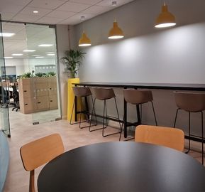 A modern workplace lounge area featuring a high counter with brown bar stools under yellow pendant lights, next to a curved glass-block wall and a round meeting table.