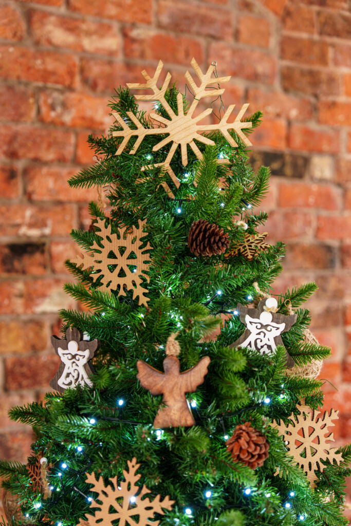 A decorated Christmas tree with wooden snowflake and angel ornaments, pinecones, and white string lights, set against a rustic brick wall background