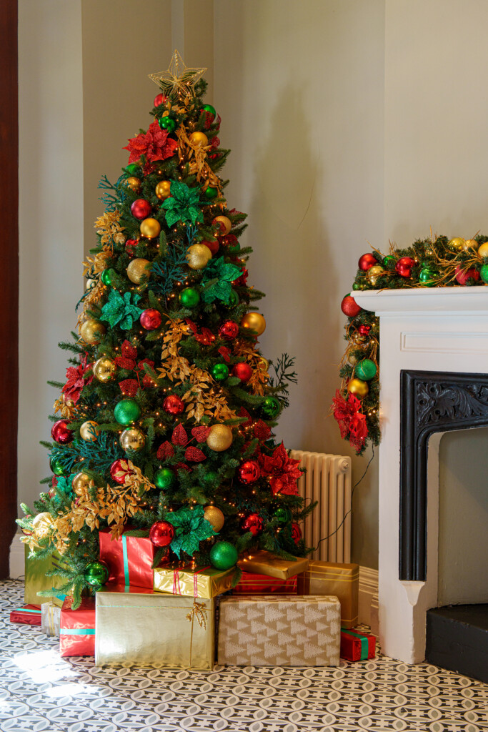 A decorated Christmas tree with ornaments stands next to a white fireplace. Wrapped gifts sit under the tree, and garland decorates the mantel.’