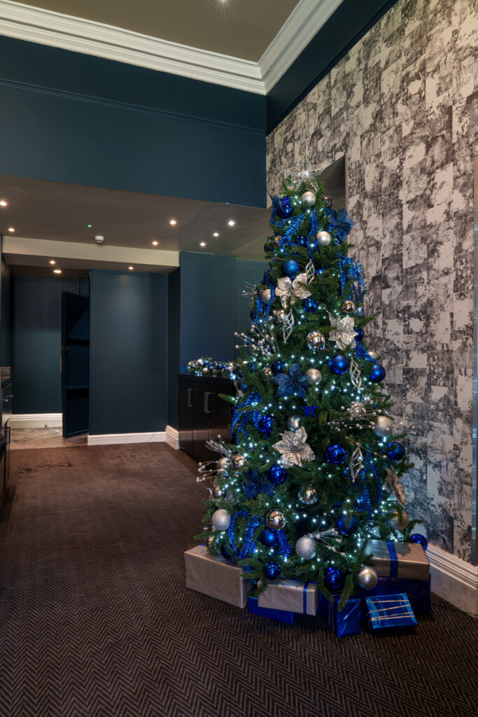 A Christmas tree decorated with blue, silver, and white ornaments stands in a modern room with dark walls and patterned wallpaper. Wrapped presents are arranged at the base of the tree.