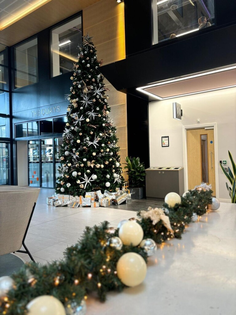 A tall Christmas tree decorated with ornaments stands in a modern building lobby. Wrapped gifts are under the tree, and a garland with white balls and silver accents decorates a nearby counter.